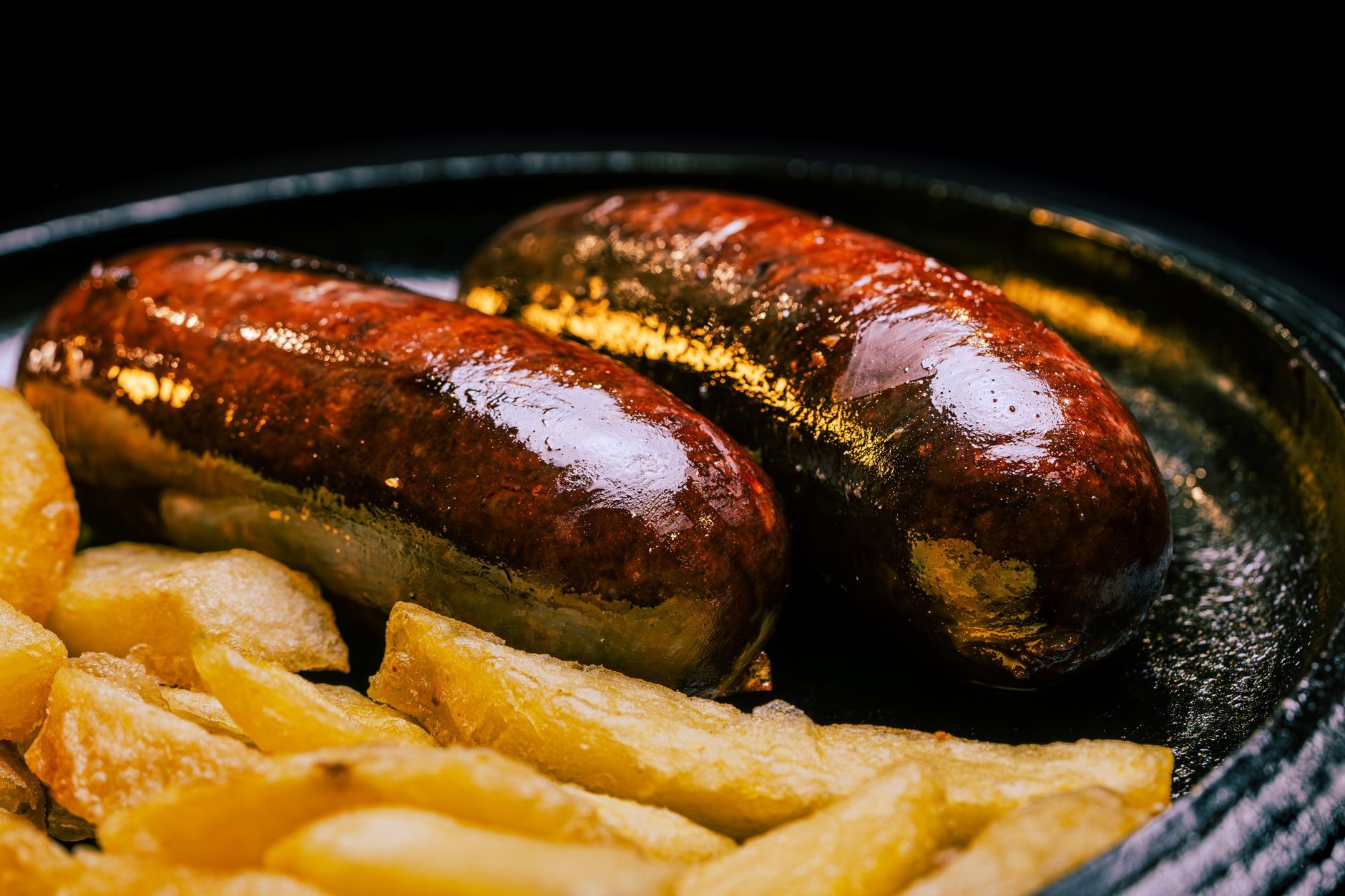 Dos piezas de morcilla tradicional asada y brillante servidas en un plato de hierro fundido caliente, acompañadas de patatas fritas caseras crujientes de fondo