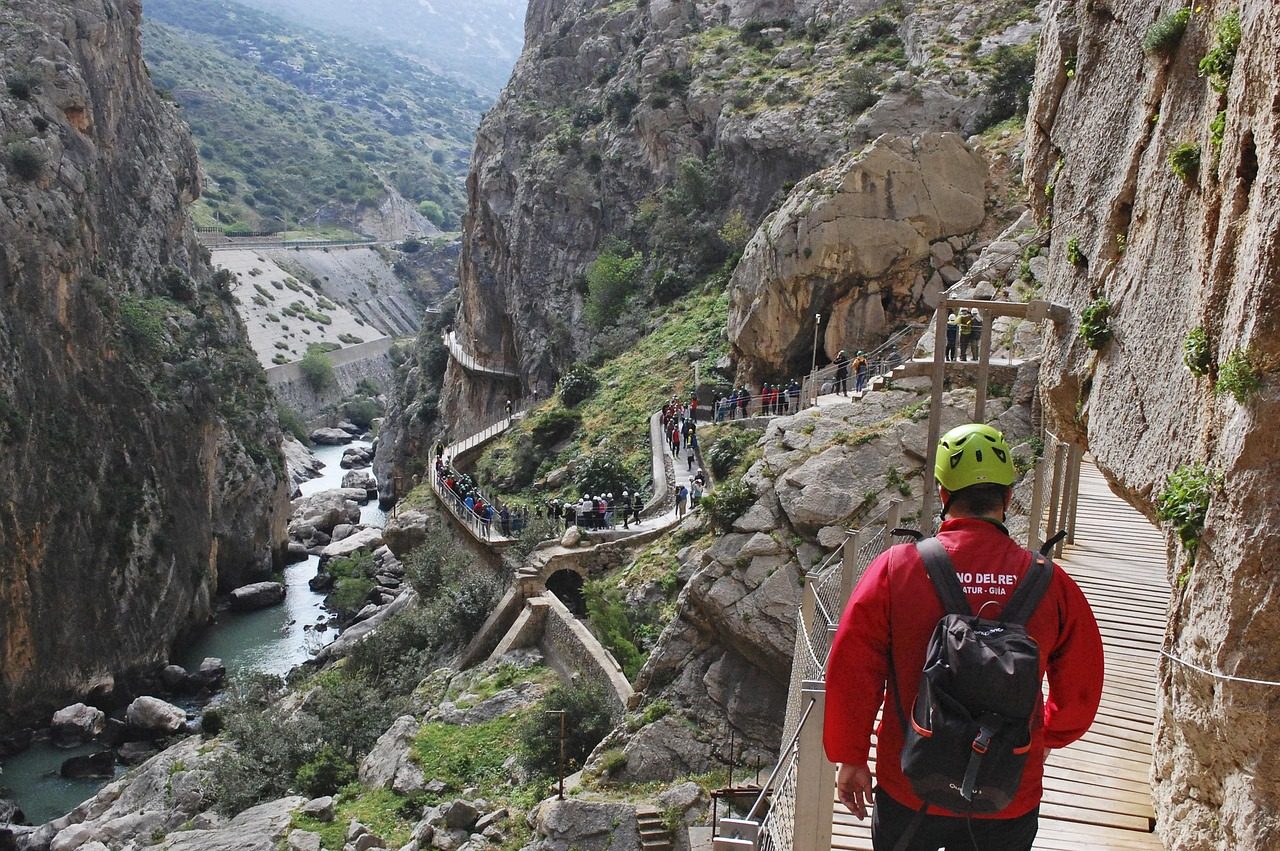 excursión organizada al Caminito del Rey en Málaga. En primer plano, un senderista equipado con mochila y casco de seguridad recorre las impresionantes pasarelas de madera colgadas del cañón rocoso, con vistas profundas al desfiladero y al río Guadalhorce