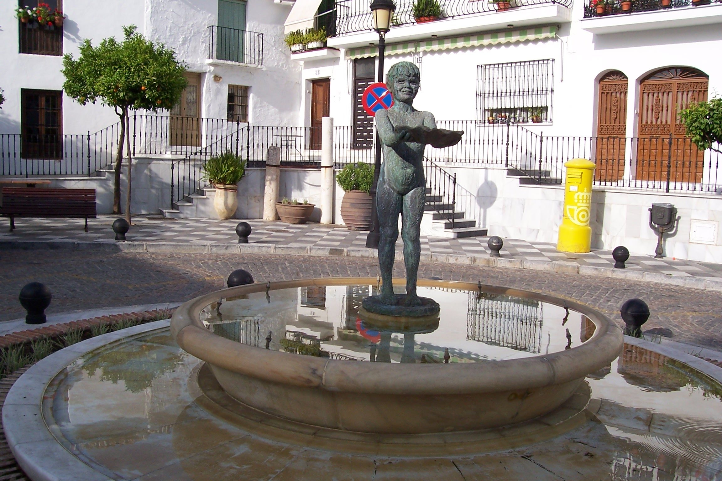 Estatua de bronce de La Niña de Benalmádena en el centro de una fuente circular de piedra, ubicada en la Plaza de España de Benalmádena Pueblo, rodeada de edificios blancos tradicionales.