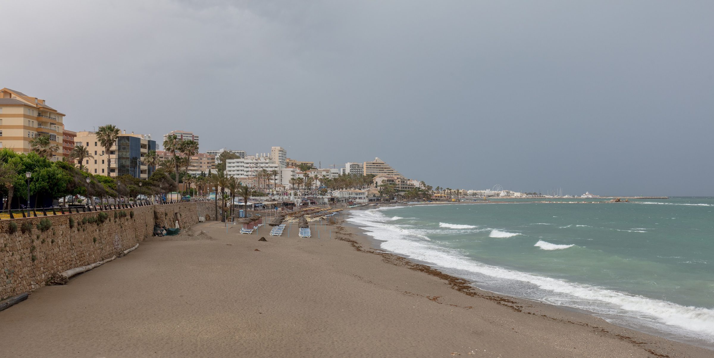 Amplia vista panorámica de una playa de arena oscura en Benalmádena en un día nublado, con edificios frente al mar al fondo