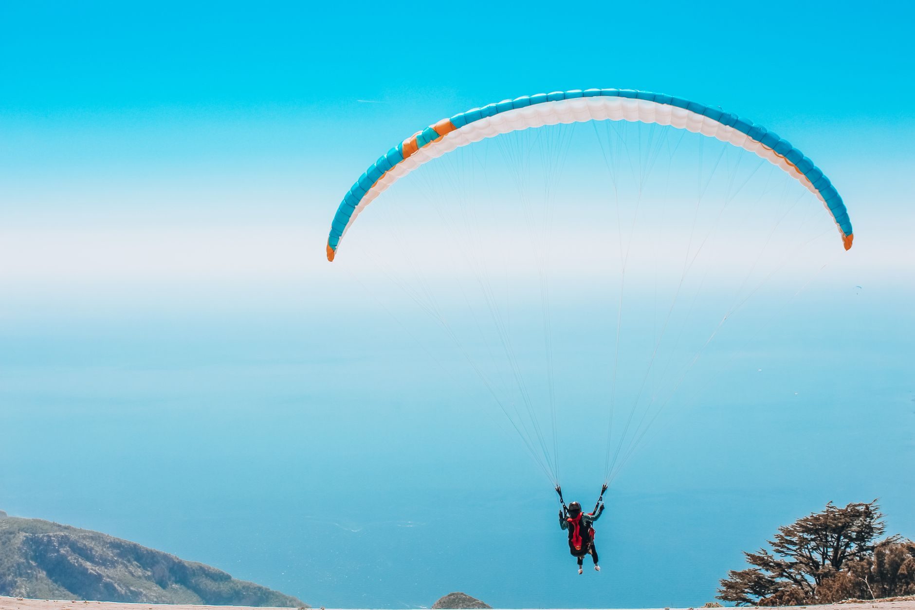 Persona volando en un parapente azul y blanco sobre un paisaje montañoso con el mar y el cielo azul de fondo