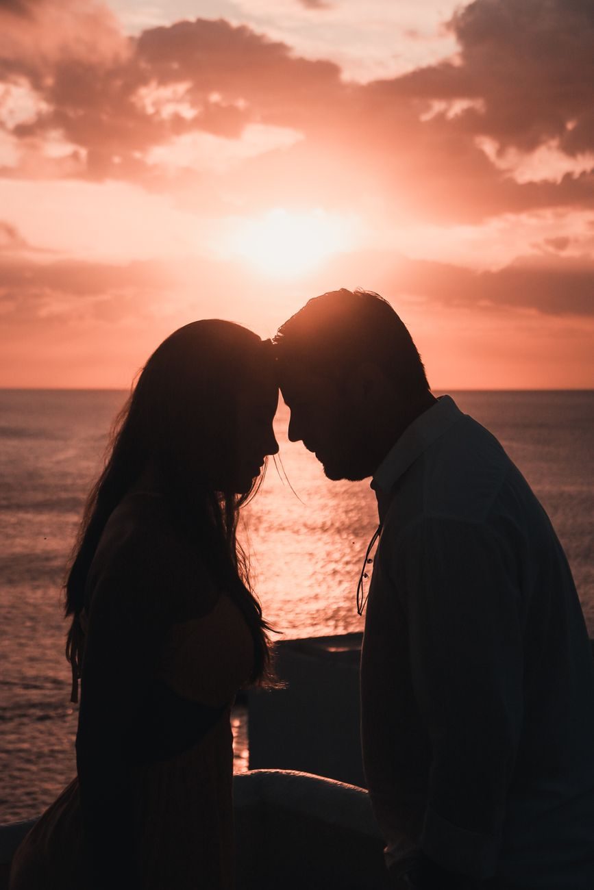 Primer plano de la silueta de un hombre y una mujer frente a frente con las frentes juntas. Al fondo se ve el mar y un cielo iluminado por los tonos naranjas y dorados de la puesta de sol.