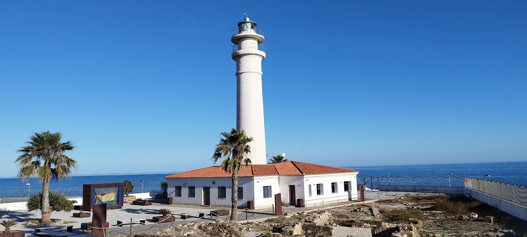 Faro de Torrox de color blanco y forma cilíndrica alzado sobre una vivienda de base cuadrada con tejados rojizos, rodeado por una palmera y muros de piedra bajo un cielo azul despejado