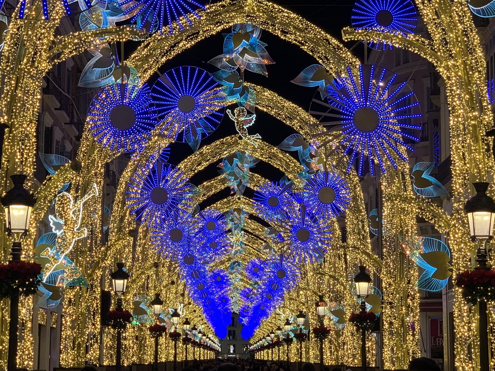 Espectacular iluminación navideña de la Calle Larios en Málaga por la noche, mostrando una majestuosa bóveda de luces doradas en arco con rosetones azules brillantes y figuras de ángeles suspendidas sobre la calle peatonal