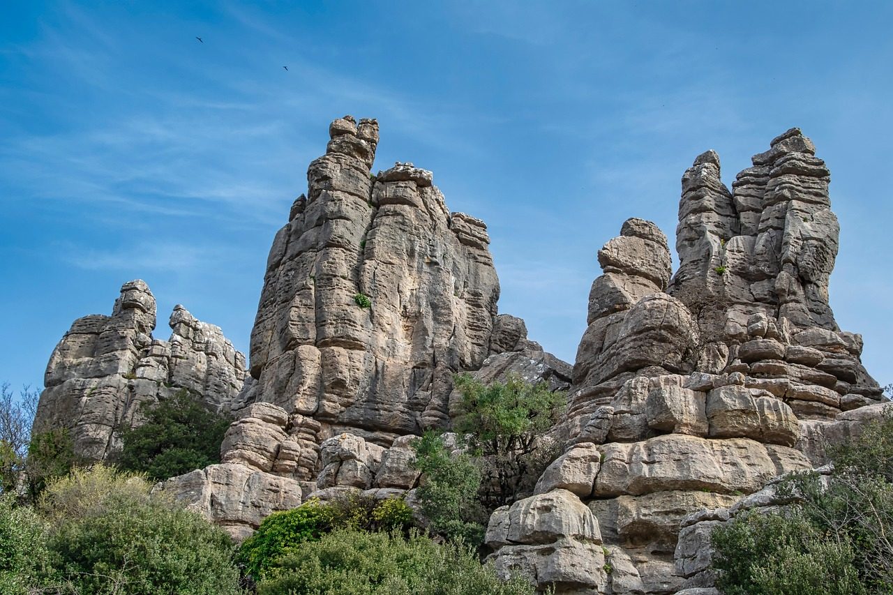 Imponentes torres y columnas de piedra caliza erosionada en el Torcal de Antequera, rodeadas de vegetación baja mediterránea bajo un cielo azul con nubes ligeras.