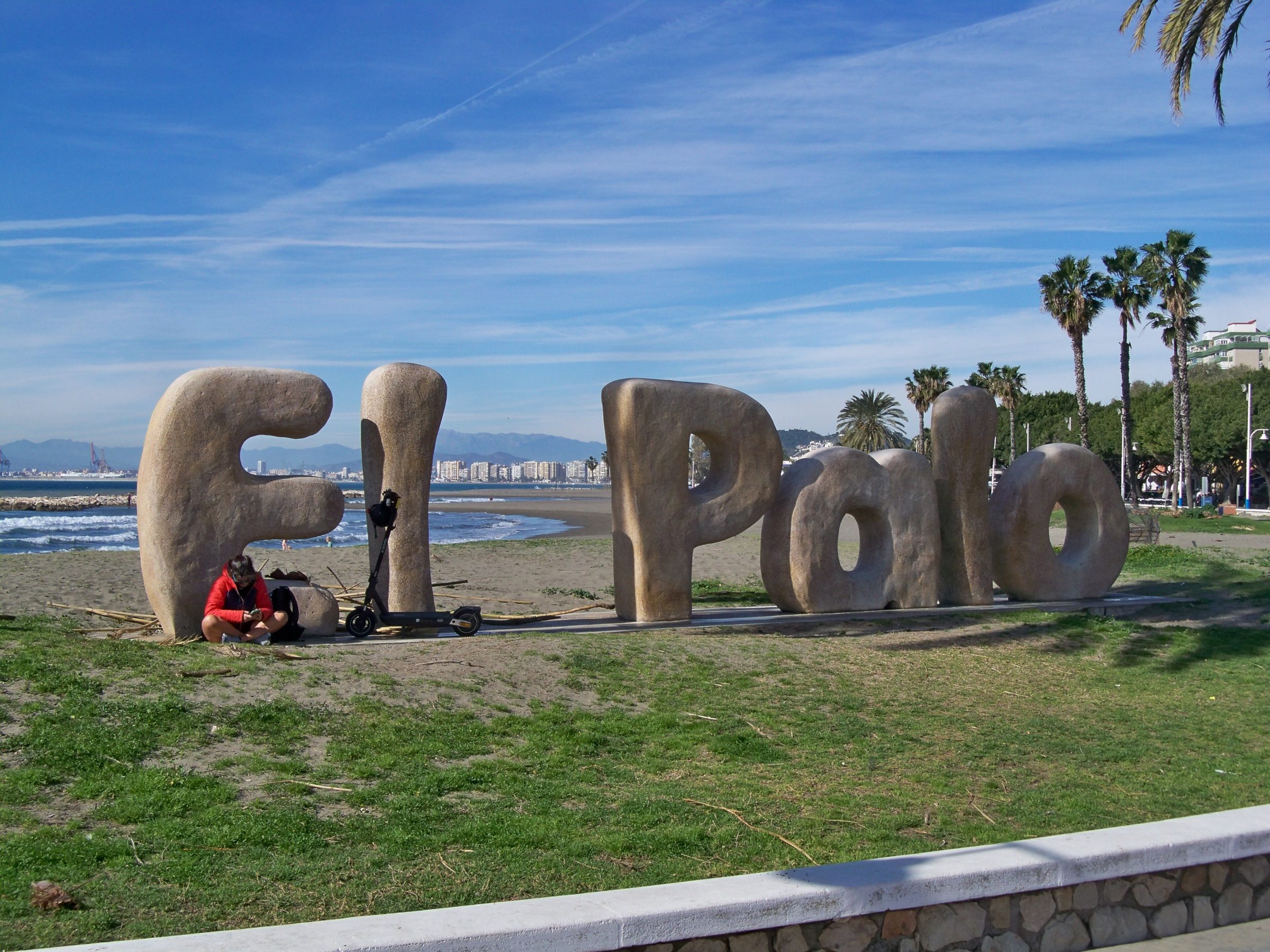 Grandes letras de piedra con la inscripción 'El Palo' situadas en la playa del Palo en Málaga, con el mar, palmeras y el skyline de la ciudad al fondo en un día soleado de invierno.