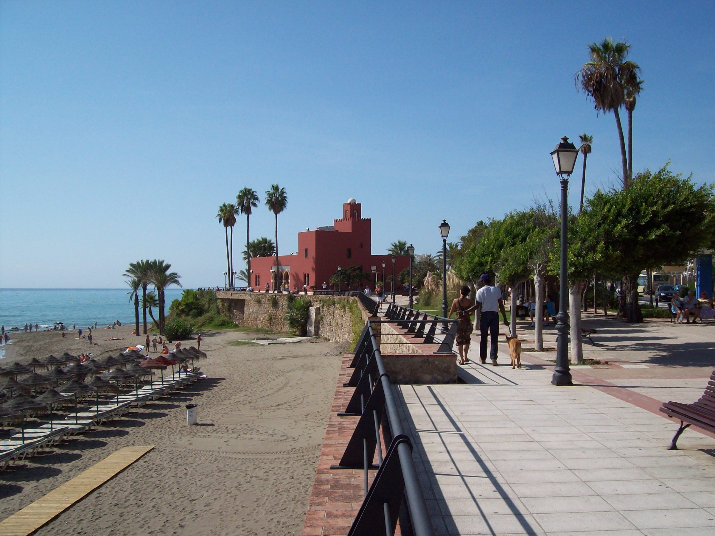 Paseo marítimo peatonal junto a la playa de Benalmádena y el Castillo de Bil Bil de color rojo al fondo frente al mar Mediterráneo