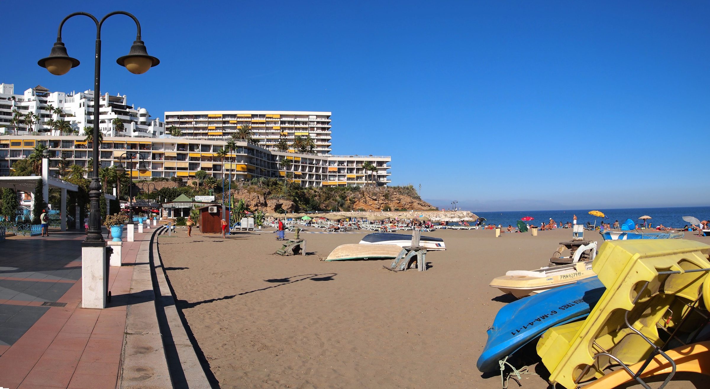 Paseo marítimo adoquinado de la Playa del Bajondillo en Torremolinos, con barcas de pedales en la arena y un gran edificio de apartamentos sobre el acantilado.